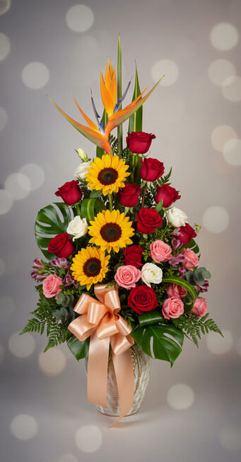 Arreglo floral alto en jarrón de vidrio con girasoles amarillos, rosas rojas, rosas rosadas y blancas, hojas verdes grandes, lazos color durazno y flores ave del paraíso sobre fondo desenfocado claro
