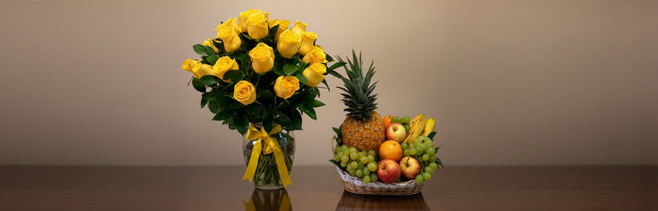 Jarrón de vidrio con ramo de rosas amarillas y lazo amarillo junto a frutero de mimbre con piña, uvas, manzanas, naranjas y plátanos sobre mesa oscura y fondo beige liso