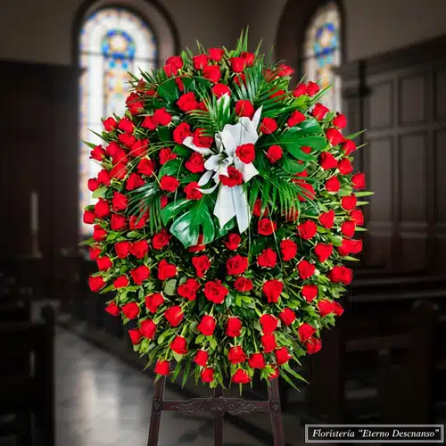 Arreglo floral funerario ovalado con numerosas rosas rojas y follaje verde, lazo blanco al centro, colocado sobre caballete de madera dentro de una iglesia