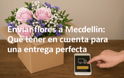 Ramo de rosas rosadas y flores blancas y moradas dentro de una caja de cartón sobre una mesa de madera, junto a una mano que toca la pantalla de un teléfono móvil con icono de camión de reparto y texto superpuesto que dice 'Enviar flores a Medellín: Qué tener en cuenta para una entrega perfecta'
