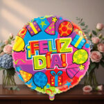 Globo redondo de colores brillantes con texto ¡Feliz día! y dibujos de regalos, globos y sombreros de fiesta, frente a un jarrón con ramos de flores sobre una mesa de madera
