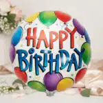 Globo redondo metálico con texto Happy Birthday y globos de colores alrededor sobre fondo decorado con flores