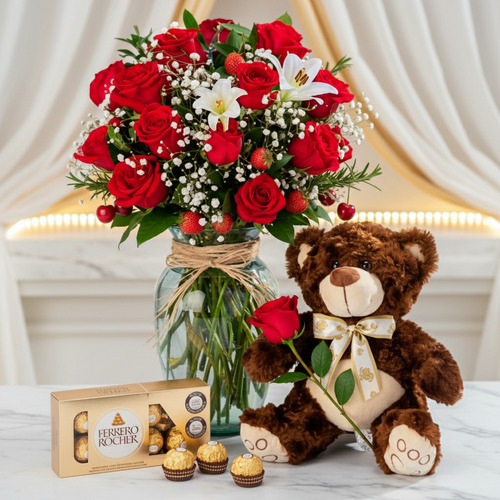 Ramo de rosas rojas y lirios blancos en jarrón de vidrio junto a oso de peluche marrón con rosa y caja de chocolates Ferrero Rocher sobre mesa de mármol