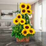 Arreglo floral vertical de girasoles amarillos con follaje verde en base rectangular de madera sobre mesa de mármol en sala moderna