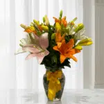 Arreglo de lirios naranjas, amarillos, blancos y rosados en un jarrón de vidrio con lazo amarillo sobre una mesa de mármol