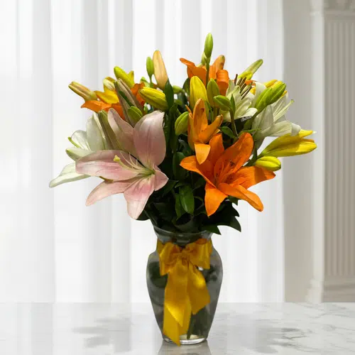 Arreglo de lirios naranjas, amarillos, blancos y rosados en un jarrón de vidrio con lazo amarillo sobre una mesa de mármol