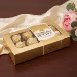 Caja dorada de bombones envueltos en papel oro sobre mesa de madera, junto a tela beige satinada y ramo de flores rosas y blancas