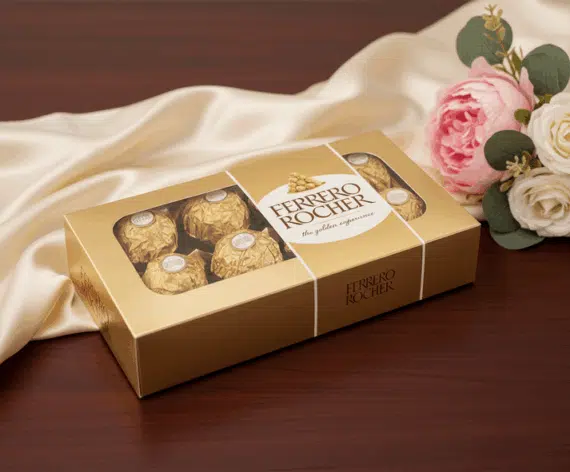 Caja dorada de bombones envueltos en papel oro sobre mesa de madera, junto a tela beige satinada y ramo de flores rosas y blancas