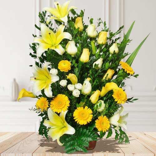 Arreglo floral vertical con lirios amarillos, rosas blancas, gerberas amarillas y follaje verde sobre una mesa de madera clara, con fondo interior desenfocado