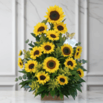 Arreglo triangular de girasoles amarillos con centro oscuro, hojas verdes y follaje, colocado en una base de madera frente a una pared de mármol claro