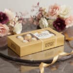 Caja dorada de bombones Ferrero Rocher sobre bandeja redonda de mármol con flores decorativas desenfocadas al fondo y cinta dorada