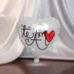 Globo plateado en forma de corazón con la frase te amo y un corazón rojo dibujado, sobre fondo de tela blanca difuminada