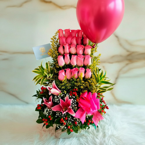 Arreglo floral vertical con rosas rosas, lirios, follaje verde, lazo rosa y globo metálico rosa sobre fondo claro