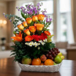 Arreglo floral con rosas naranjas, lirio naranja, margaritas y uvas, manzanas y naranjas en canasta blanca sobre mesa de madera en interior luminoso