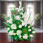 Arreglo floral de lirios blancos, rosas blancas y hortensias con follaje verde sobre una mesa de madera, con un fondo desenfocado de ventanales y más flores