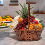 Canasta de mimbre con piña, uvas, duraznos, naranjas, kiwi, plátanos y flores blancas sobre una encimera de mármol en una cocina moderna