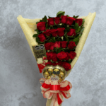 Arreglo de ramo envuelto en papel crema con rosas rojas en varias filas, chocolates Ferrero Rocher en la base y barra de chocolate Mont Blanc al lado, decorado con moño rojo y crema sobre fondo gris marmoleado