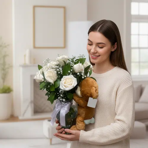 Mujer sonriente con suéter beige sostiene un ramo de rosas blancas y gypsophila en jarrón de vidrio decorado con lazo lila y un osito de peluche marrón en una sala luminosa