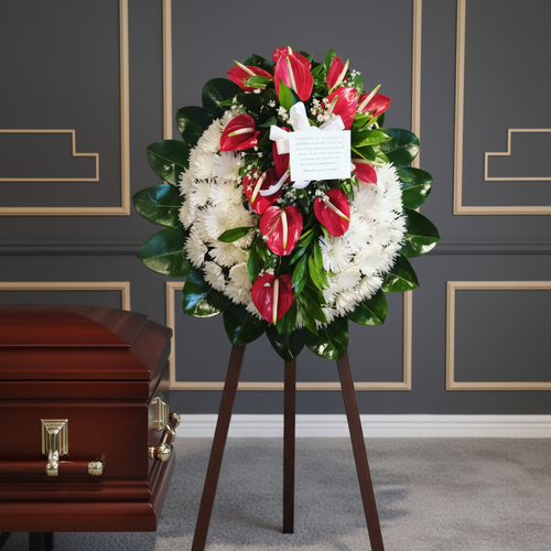 Corona funeraria redonda con flores blancas y lirios rojos sobre caballete de madera, colocada junto a un ataúd de madera brillante en una sala con paredes grises decoradas con molduras doradas