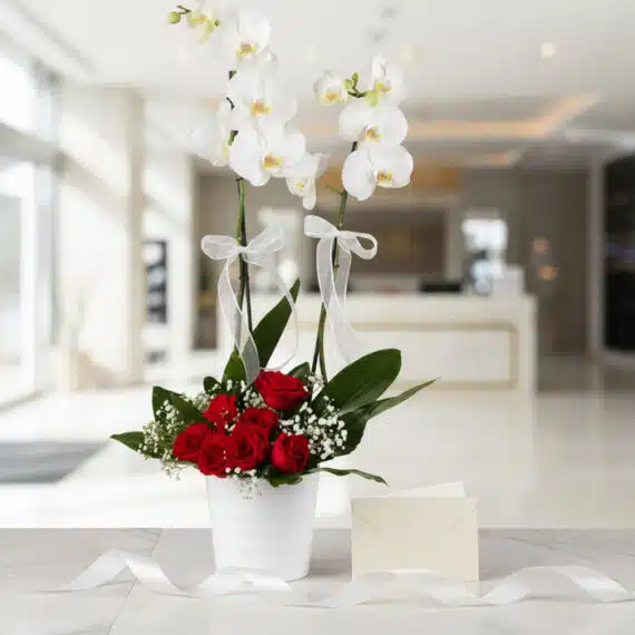 Arreglo floral BRIANA con orquídeas blancas y rosas rojas, tarjeta de regalo y cinta satinada en un elegante lobby.