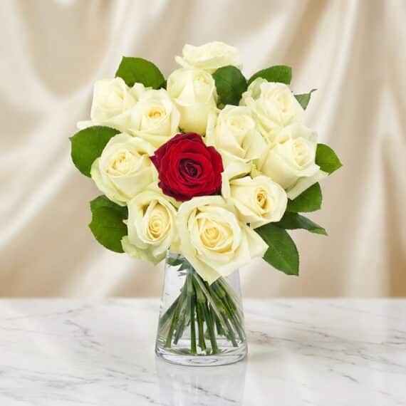 Arreglo floral CARLA con 12 rosas de tallo largo, una roja central y once blancas, en un jarrón de cristal sobre mármol.