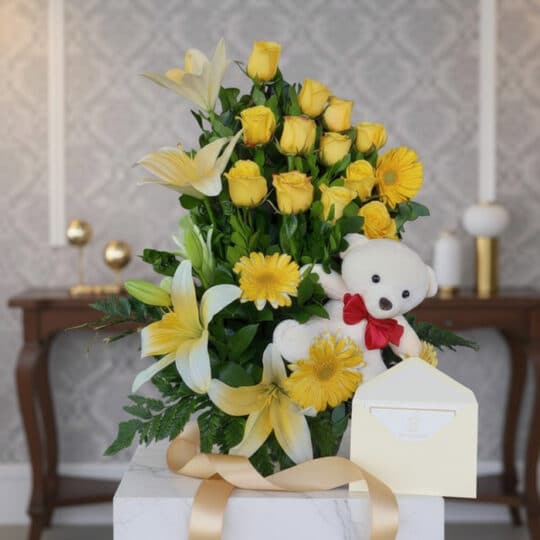 Arreglo floral exótico Amaretto con rosas amarillas, lirios y gerberas, acompañado de un oso de peluche blanco y cinta dorada