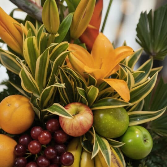 Primer plano extremo de Arreglo Floral con Frutas Bahamas, mostrando lirios naranjas, hojas verdes variegadas, naranjas, uvas