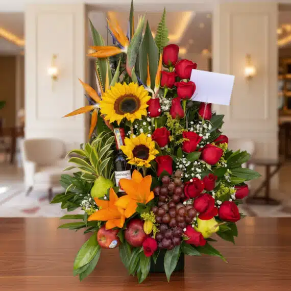 Arreglo floral premium con girasoles, rosas rojas, lirios, uvas, manzanas, peras y una botella de vino, sobre una mesa de mad