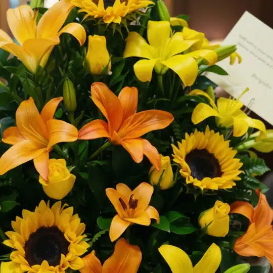 Primer plano extremo de un arreglo floral tropical con lirios naranjas, girasoles amarillos y rosas amarillas, destacando tex