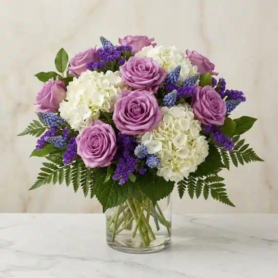 Arreglo floral VITTORIA con rosas lilas, hortensias blancas y muscari azul en un jarrón de cristal, sobre una mesa de mármol.