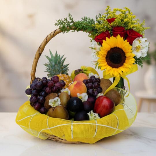 Canasta de Frutas Draco con uvas, manzanas, duraznos, kiwis, piña y arreglo floral de rosas, girasol y flores blancas sobre m