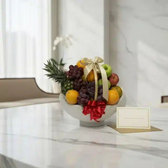 Canasta de frutas gourmet con piña, uvas, manzanas y naranjas, decorada con cintas doradas y rojas, sobre una mesa de mármol