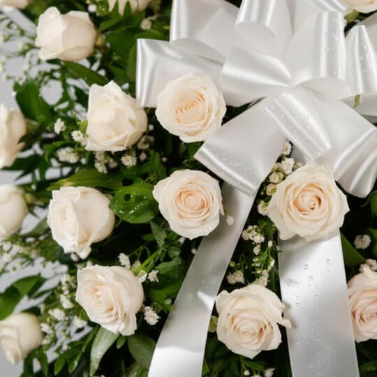 Fotografía macro premium de la Corona Fúnebre Serenidad, mostrando rosas blancas con rocío, follaje verde y un lazo de satén