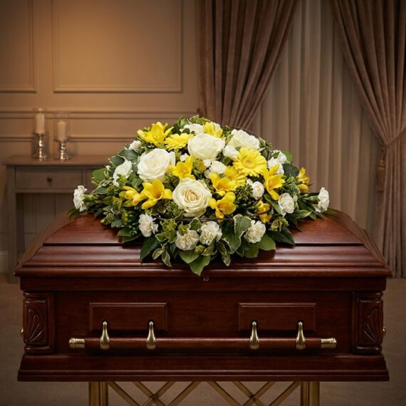 Cubre Caja Fúnebre Baltasar: Homenaje Floral de Serenidad y Respeto Cubre Caja Fúnebre Baltasar con rosas blancas, freesias amarillas y margaritas, sobre un ataúd de madera oscura en un entorno