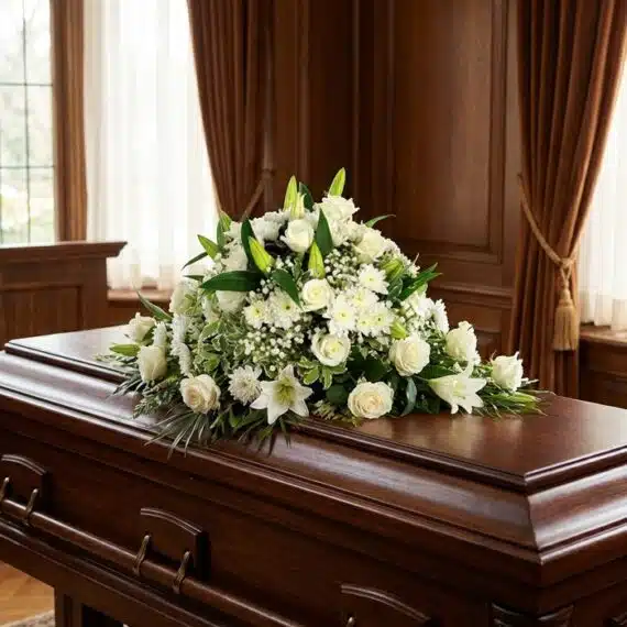 Cubre Caja Fúnebre Solemne: El Respetuoso Homenaje a Lidia 🕊️ Cubre caja fúnebre solemne con rosas blancas, lirios y follaje verde sobre un ataúd de madera oscura en un entorno elegante y