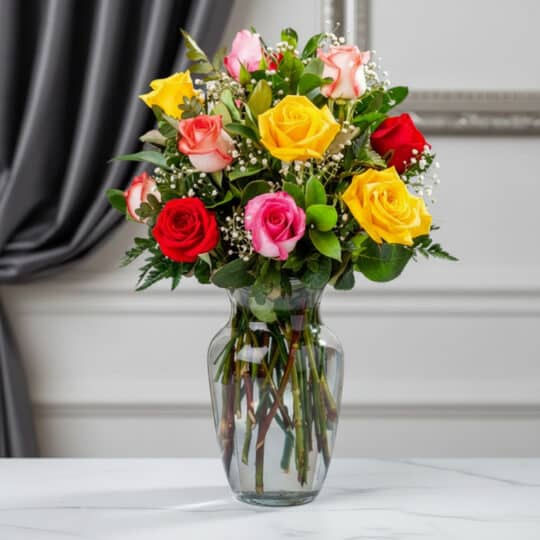 Jarrón de cristal transparente con un ramo de 12 rosas de colores variados: rojas, amarillas, rosas y blancas, con follaje ve