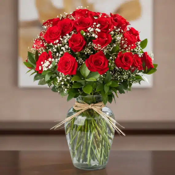 Jarrón con 24 Rosas Rojas Premium: Elegancia Floral para Regalar Jarrón de cristal con 24 rosas rojas frescas y follaje verde, atado con un lazo de rafia, sobre una mesa de madera oscura con