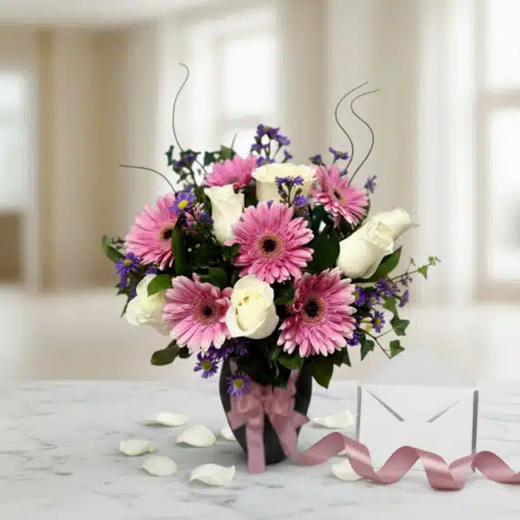Jarrón Primavera con gerberas rosas, rosas blancas y follaje, sobre mármol con tarjeta de regalo y cinta satinada, listo para