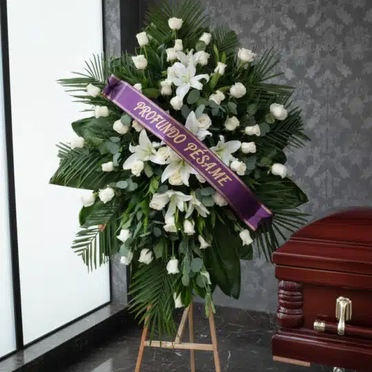 Pedestal fúnebre Eternidad con rosas blancas, lirios y follaje verde, adornado con una cinta morada que dice 'Profundo Pésame