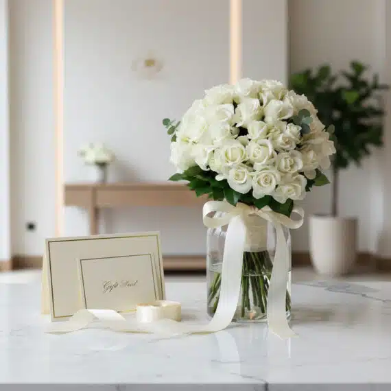 Ramo JACOBELLA de 60 rosas blancas en jarrón de cristal con cinta satinada, tarjeta de regalo y sobre sobre superficie de már