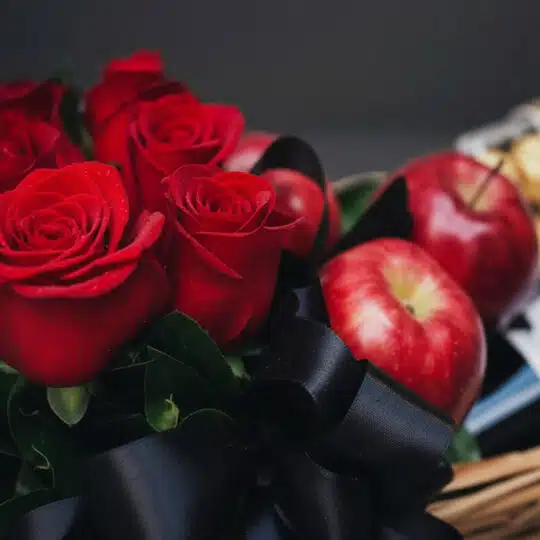 Primer plano de rosas rojas frescas con gotas de rocío, manzanas rojas brillantes y un lazo negro de satén en una cesta de re